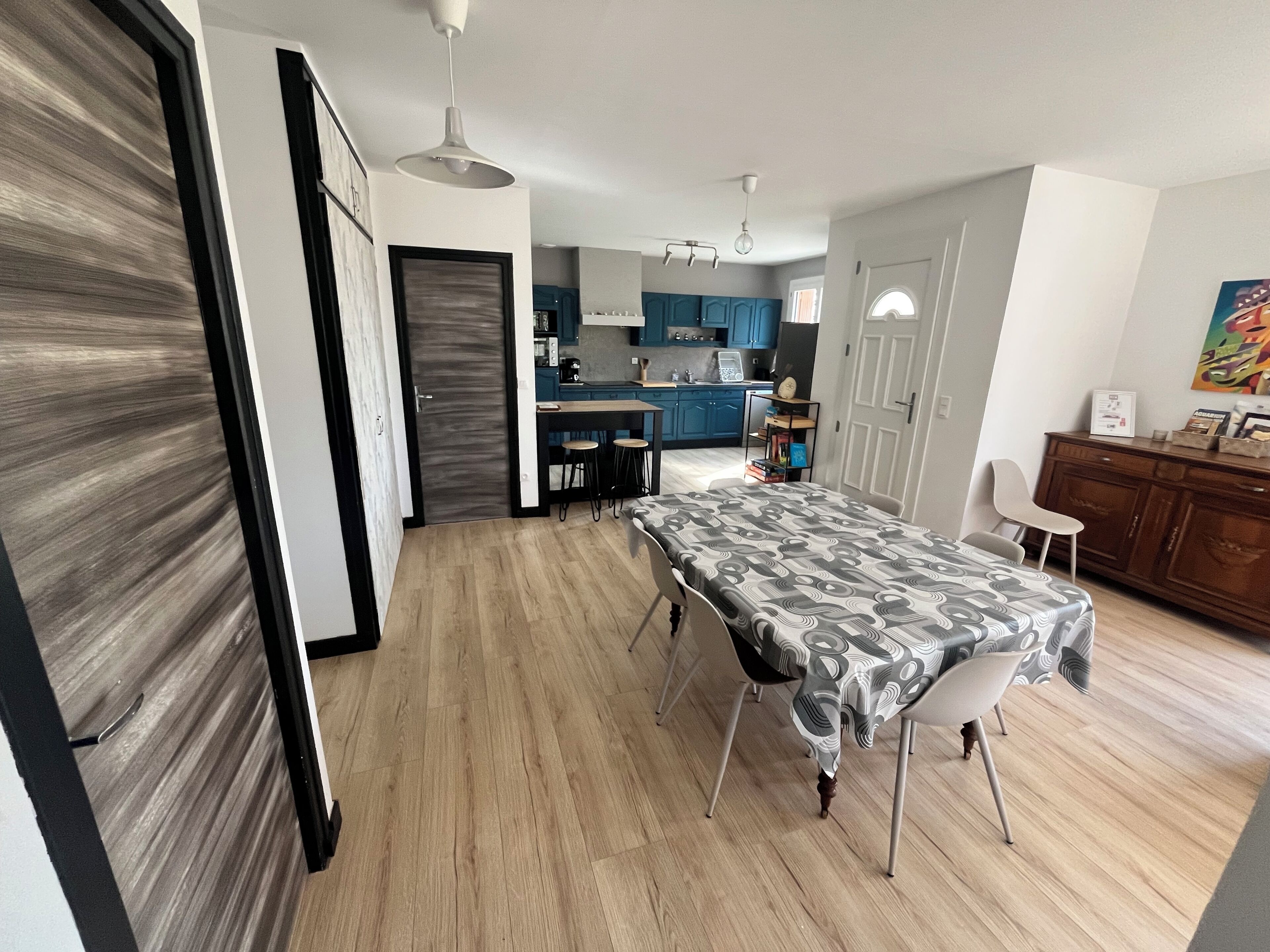 Een moderne keuken met blauwe kastjes, een eethoek met een tafelkleed met dessin, en een houten vloer.