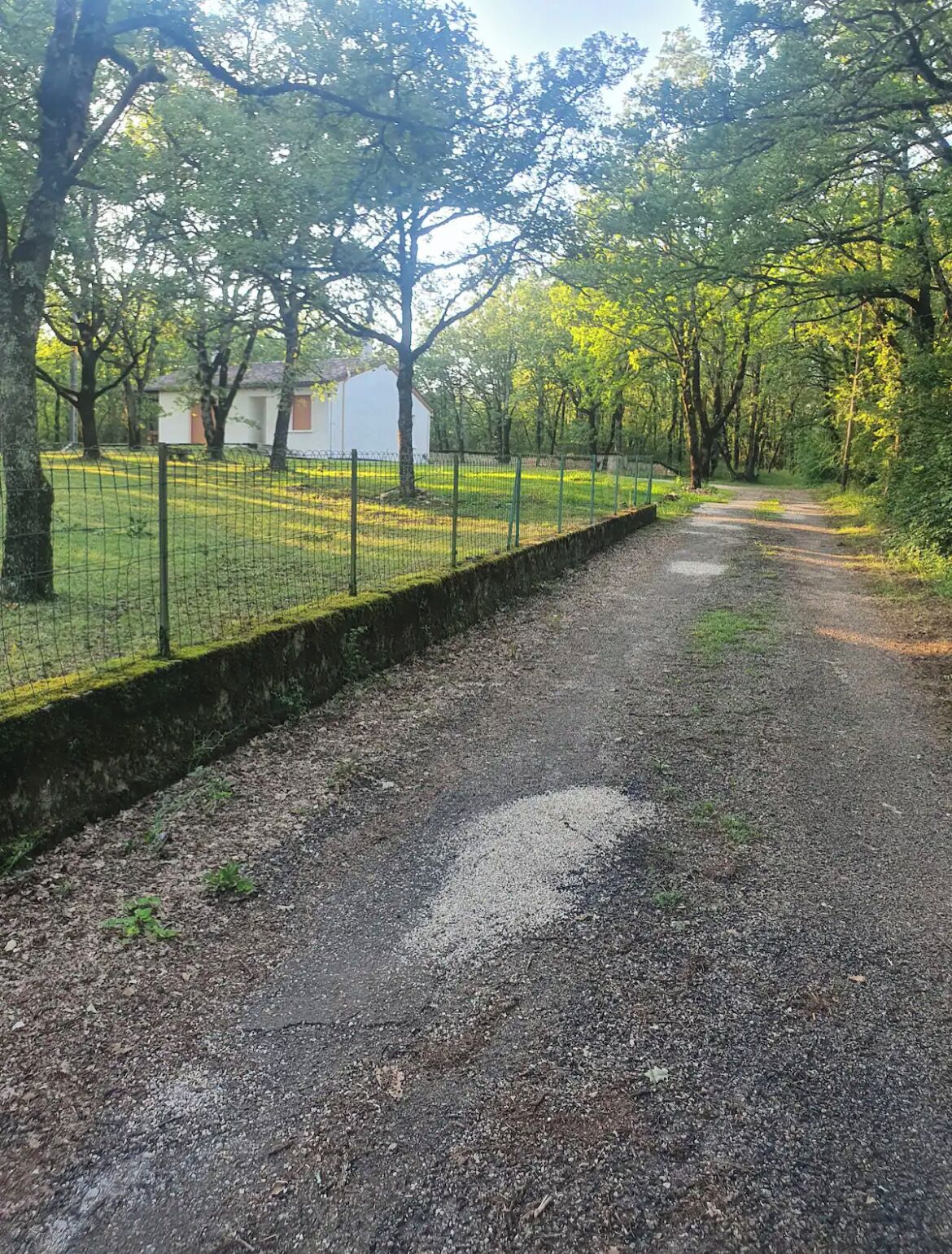 Un chemin de gravier bordé d’une clôture en fil de fer et d’arbres, qui mène à un bâtiment blanc avec une porte rouge.