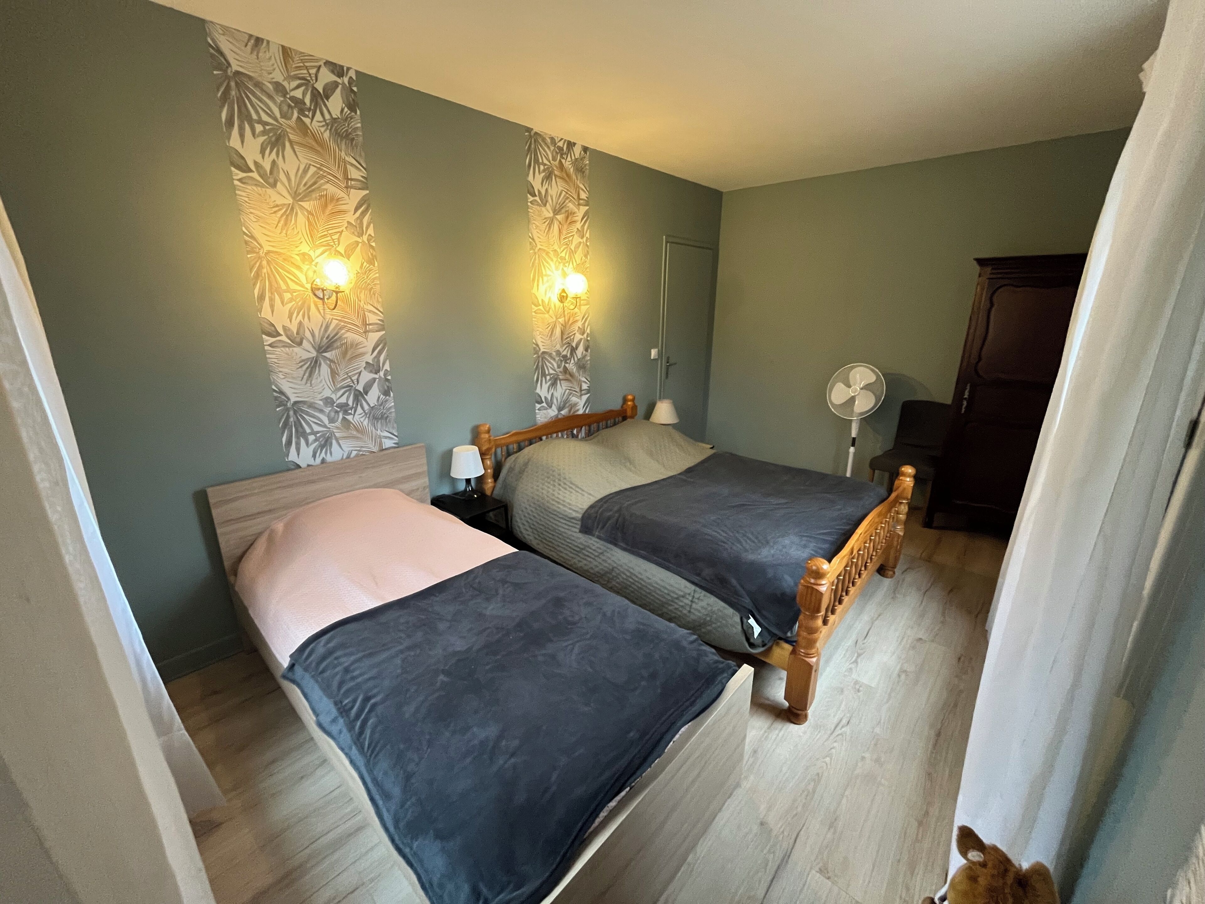 Ein Zimmer mit zwei Betten, einem Holz-Kopfstück, einem Nachttisch mit Lampe, einer Wandleuchte und einem Standventilator.