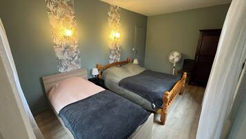Ein Zimmer mit zwei Betten, einem Holz-Kopfstück, einem Nachttisch mit Lampe, einer Wandleuchte und einem Standventilator.