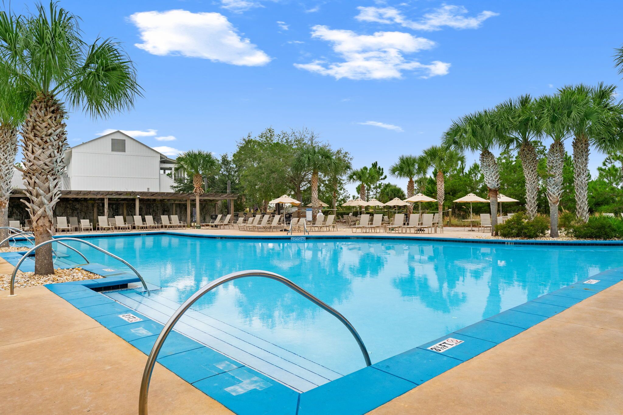 A spacious outdoor pool area features crystal-clear water, surrounded by palm trees and lounge chairs under umbrellas, offering a serene daytime retreat.