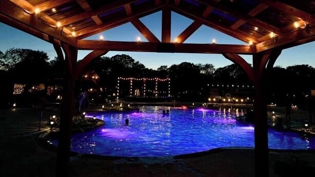Un gazebo con piscina illuminata da luci blu, immerso tra alberi e edifici.