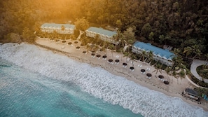 An aerial view of a beachfront hotel with multiple buildings, a sandy beach, and palm trees. - Soho Beach House Canouan (Canouan Island)