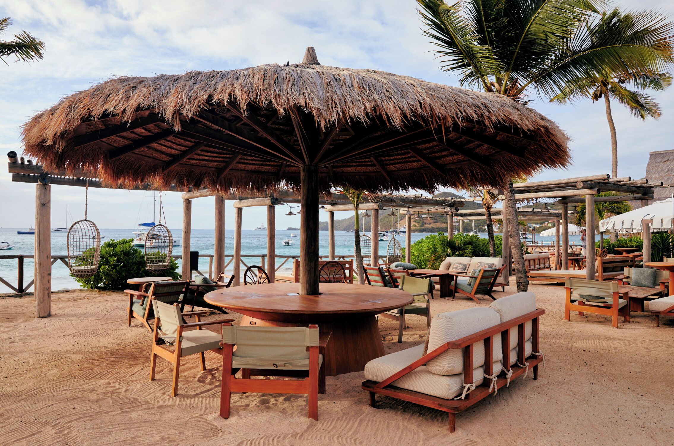 Un área de estar al aire libre frente a la playa, con sombrilla de paja, sillas y mesas de madera.