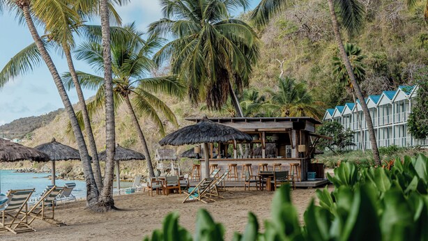 A beachside resort with thatched umbrellas, lounge chairs, and a wooden bar area. - Soho Beach House Canouan (Canouan Island)