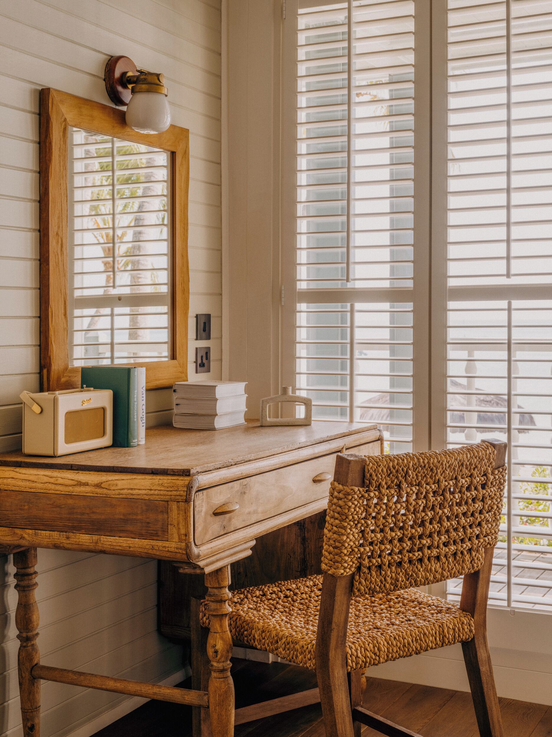 A wooden desk with a mirror, books, and a radio. A wicker chair is in front of the desk.