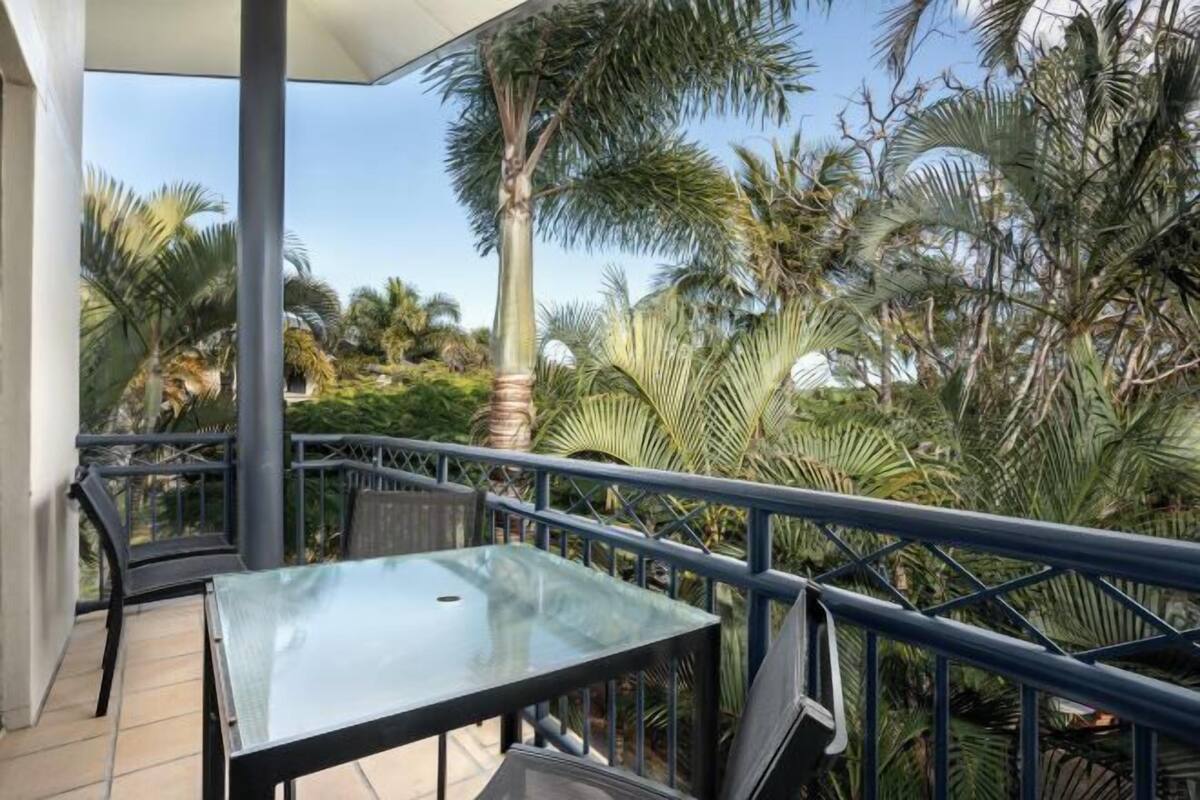 A covered outdoor area with a glass table and chairs, surrounded by palm trees and a metal railing.