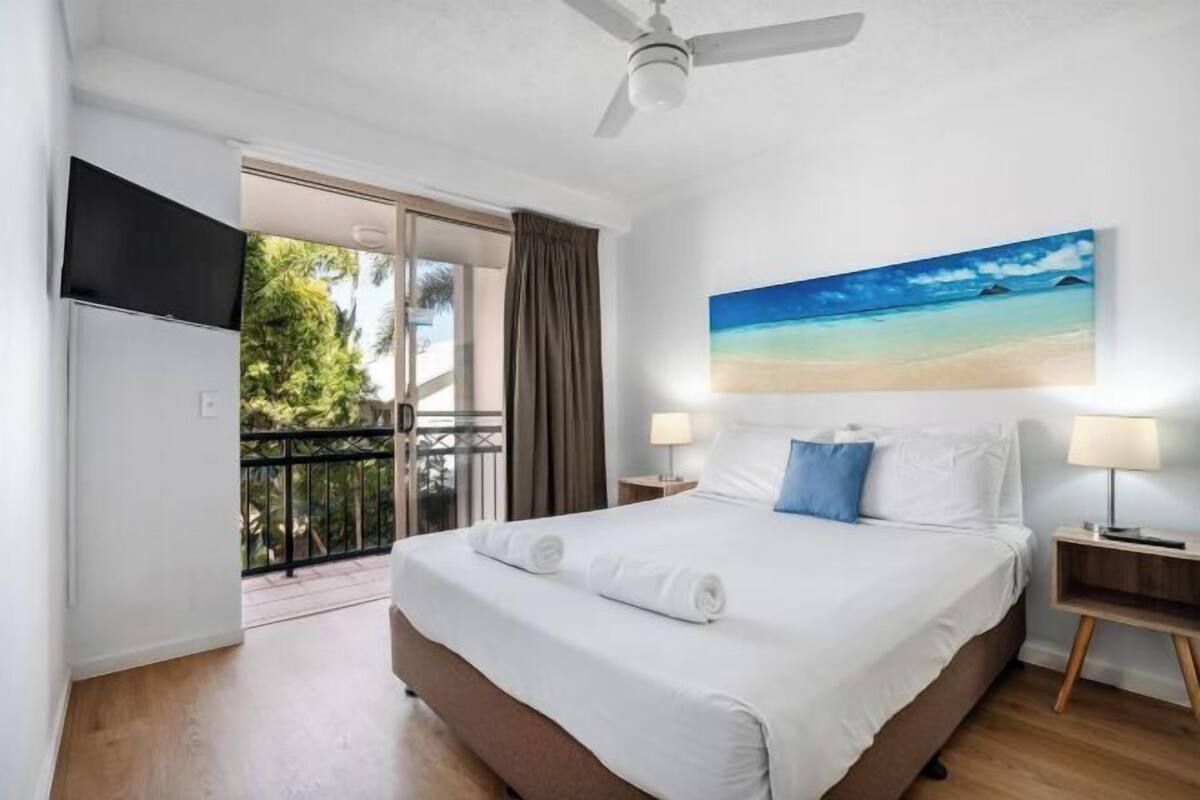 A hotel room with a bed, a painting of a beach, a ceiling fan, and a view of the outdoors.