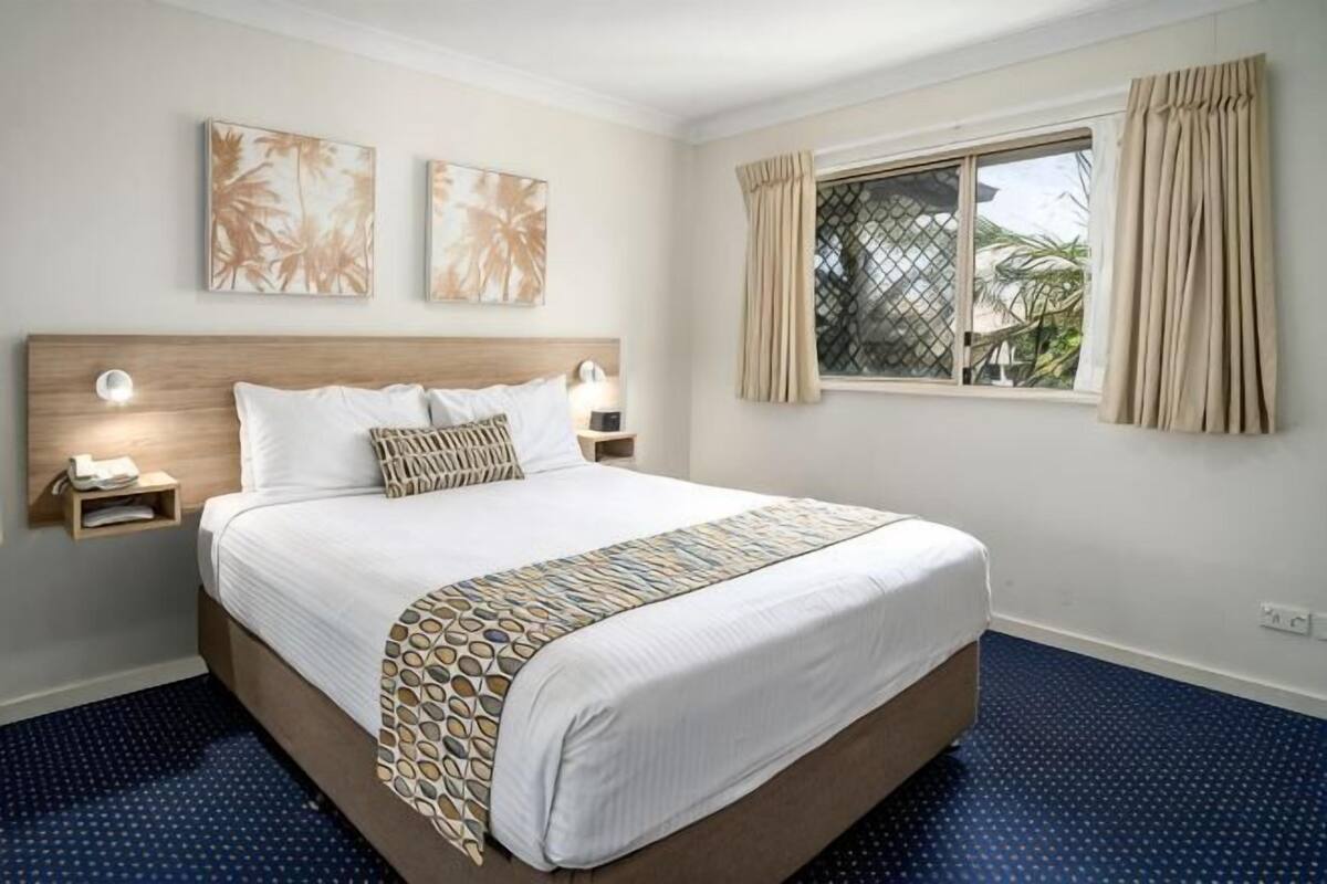 A hotel room with a large bed, two bedside tables with lamps, a window with curtains, and artwork on the wall.
