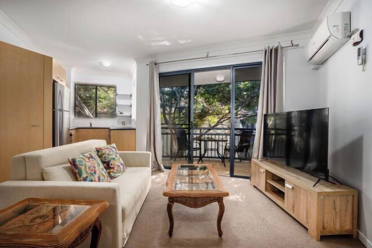 A modern living room with a sofa, a glass-top coffee table, a TV stand, and a sliding glass door leading to an outdoor area.