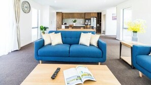 Ein modernes Wohnzimmer mit einem blauen Sofa, einem Couchtisch mit Fernbedienung und Zeitschrift, und einer Küche im Hintergrund.