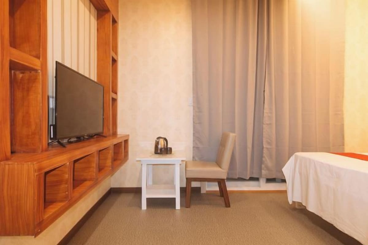 Une pièce avec un meuble TV en bois, une petite table blanche avec une bouilloire, une chaise et un lit recouvert d’un drap blanc.