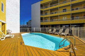 A pool area with a wooden deck, lounge chairs, and a yellow building with balconies. - Slo M'ocean - Your Stylish Beach Retreat (Carolina Beach)