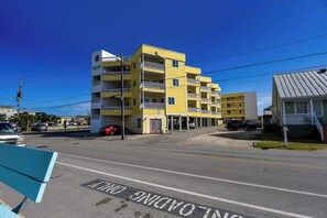 A multi-story yellow building with balconies, a parking area, and a clear blue sky. - Slo M'ocean - Your Stylish Beach Retreat (Carolina Beach)