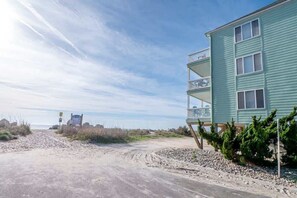 A beachfront building with a balcony, a sandy path leading to the beach, and a clear sky.