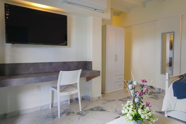 A modern hotel room with a flat-screen TV mounted on the wall, a white chair, a marble-tiled floor, a white wardrobe, and a decorative vase with flowers.