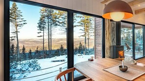 A cozy cabin interior with a large window offering a snowy forest view, a wooden dining table, and a fireplace.