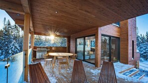 Een houten terras met een ronde tafel en stoelen, een glazen deur naar een woongedeelte en een uitzicht op besneeuwde bomen.