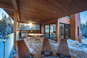 A wooden deck with a round table and chairs, a glass door leading to a living area, and a view of snow-covered trees. - RIDGEVIEW (Lac-Beauport, Quebec)