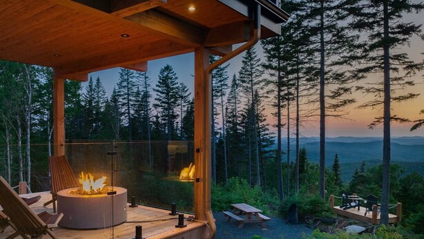 A wooden deck with a fire pit, overlooking a forested landscape with picnic tables.