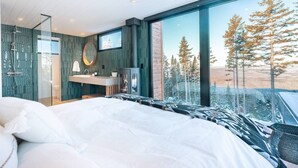 A modern bedroom with a large bed, a bathroom with a glass shower enclosure, and a view of trees through large windows.
