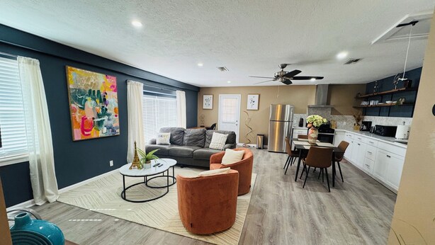 A modern living room with a grey sofa, a coffee table, and a dining area with a table and chairs. There is a kitchen area with white cabinets and a refrigerator. - Stylish Cozy 2-bedroom Duplex  in enjoyable Stafford with WiFi, AC, Smart TVs (Stafford)