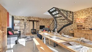A rustic interior with a stone wall, a fireplace, a wooden dining table set with plates and glasses, and a spiral staircase. - Le Tejtal 6pers - PLUMERGAT (PLUMERGAT)