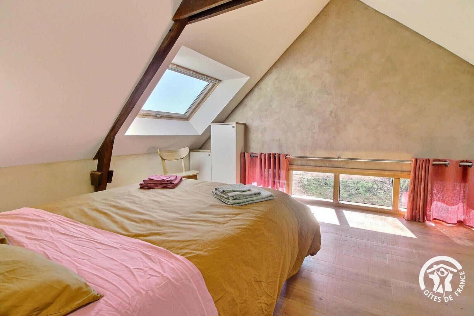 A bedroom with a sloped ceiling, a skylight, a bed with pink and yellow bedding, and a window with curtains.