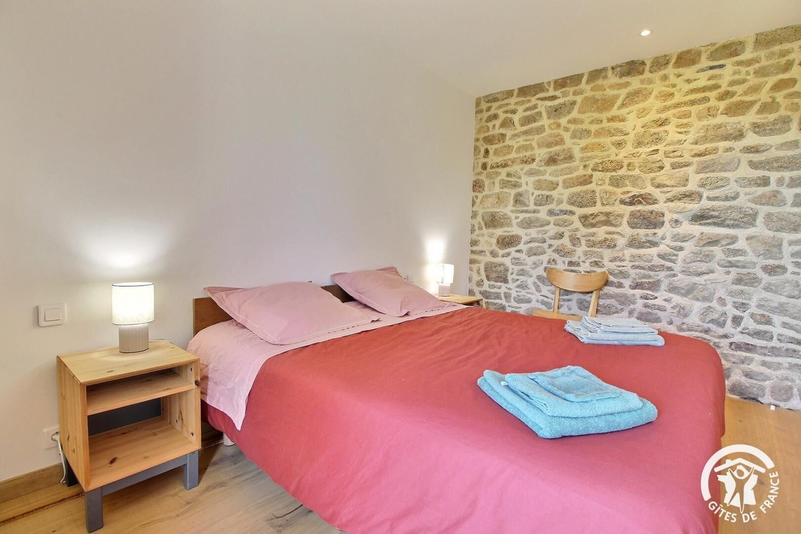 A bedroom with a stone wall, a bed with pink bedding, a wooden nightstand with a lamp, and a folded towel on the bed.