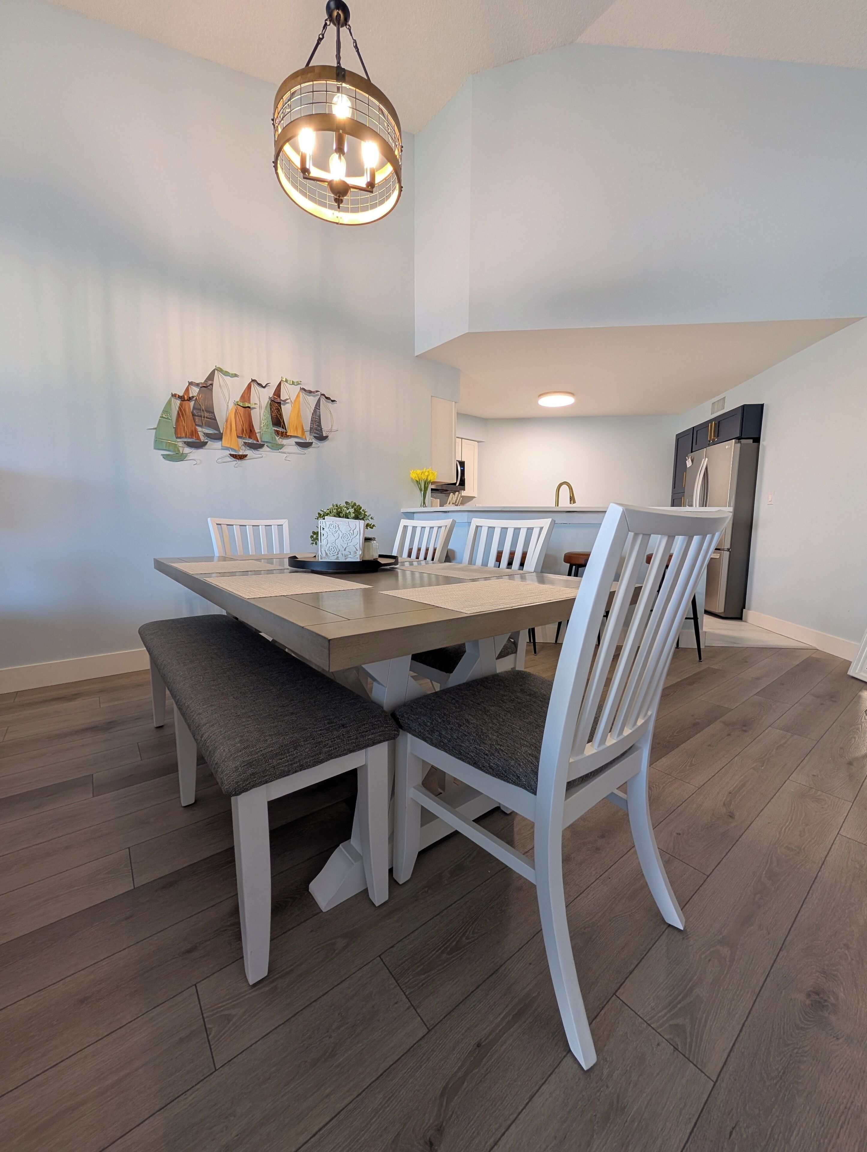 Un coin repas avec une table en bois et des chaises blanches, un lustre et une décoration murale représentant un voilier.