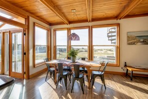 Un espace repas avec une table et des chaises en bois, de grandes fenêtres donnant sur l’extérieur et un luminaire suspendu.