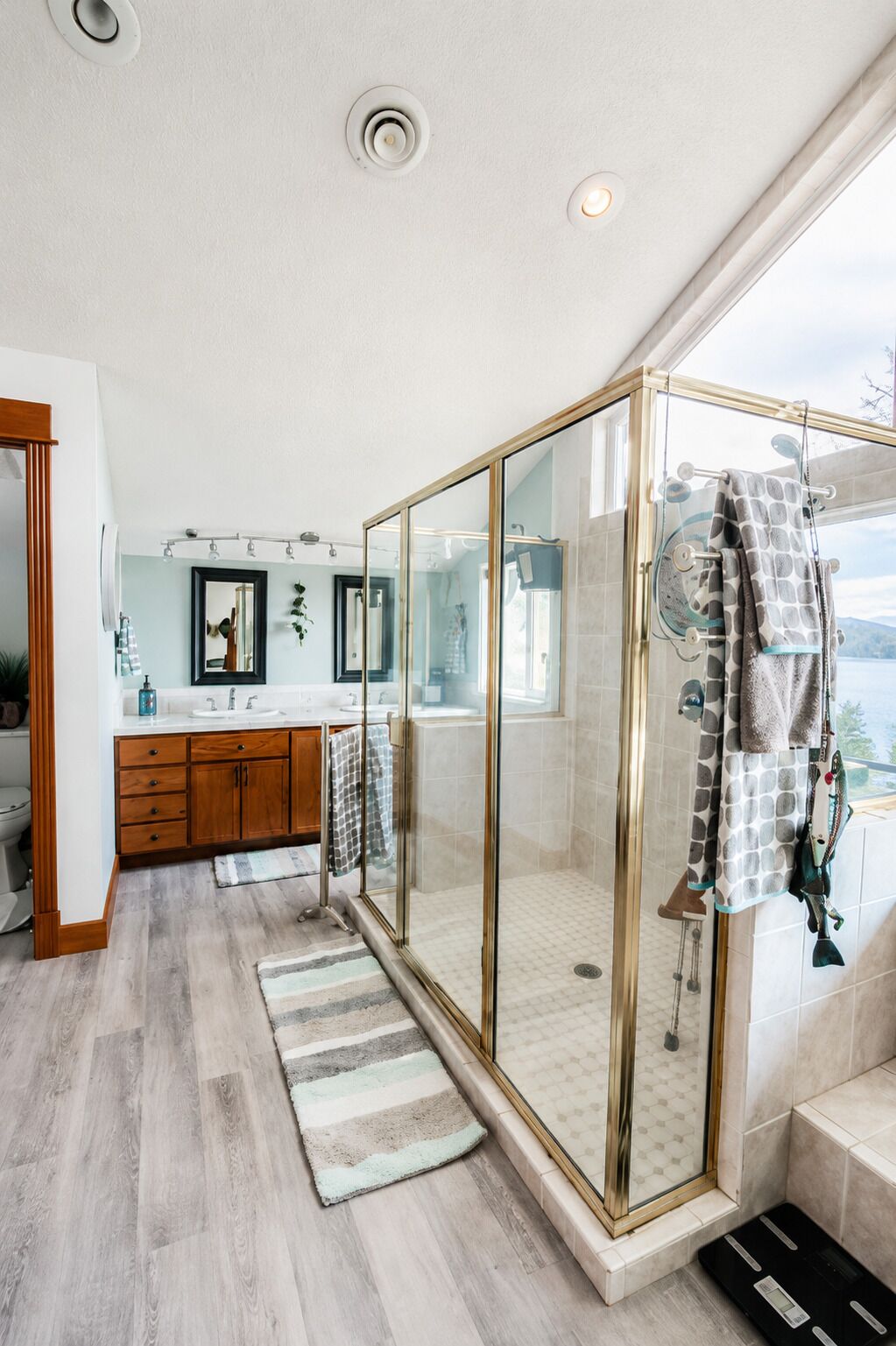 A modern bathroom with a glass-enclosed shower, wooden cabinets, a large mirror, and a view of the outdoors.