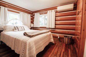 A cozy bedroom with a wooden log wall, a bed with a patterned blanket, and a window with curtains.