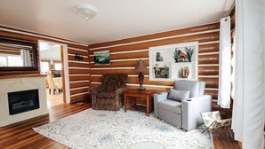 A cozy living room with a fireplace, wooden paneling, a patterned rug, and a dining area in the background. - Mayfield Lake Waterfront Escape (Mossyrock)
