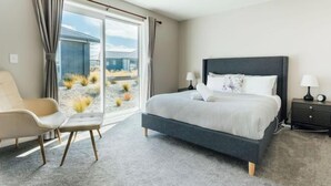 Ein modernes Schlafzimmer mit einem großen Bett, einem Sessel, einem Nachttisch und Blick auf ein Gebäude und eine Landschaft durch ein großes Fenster.