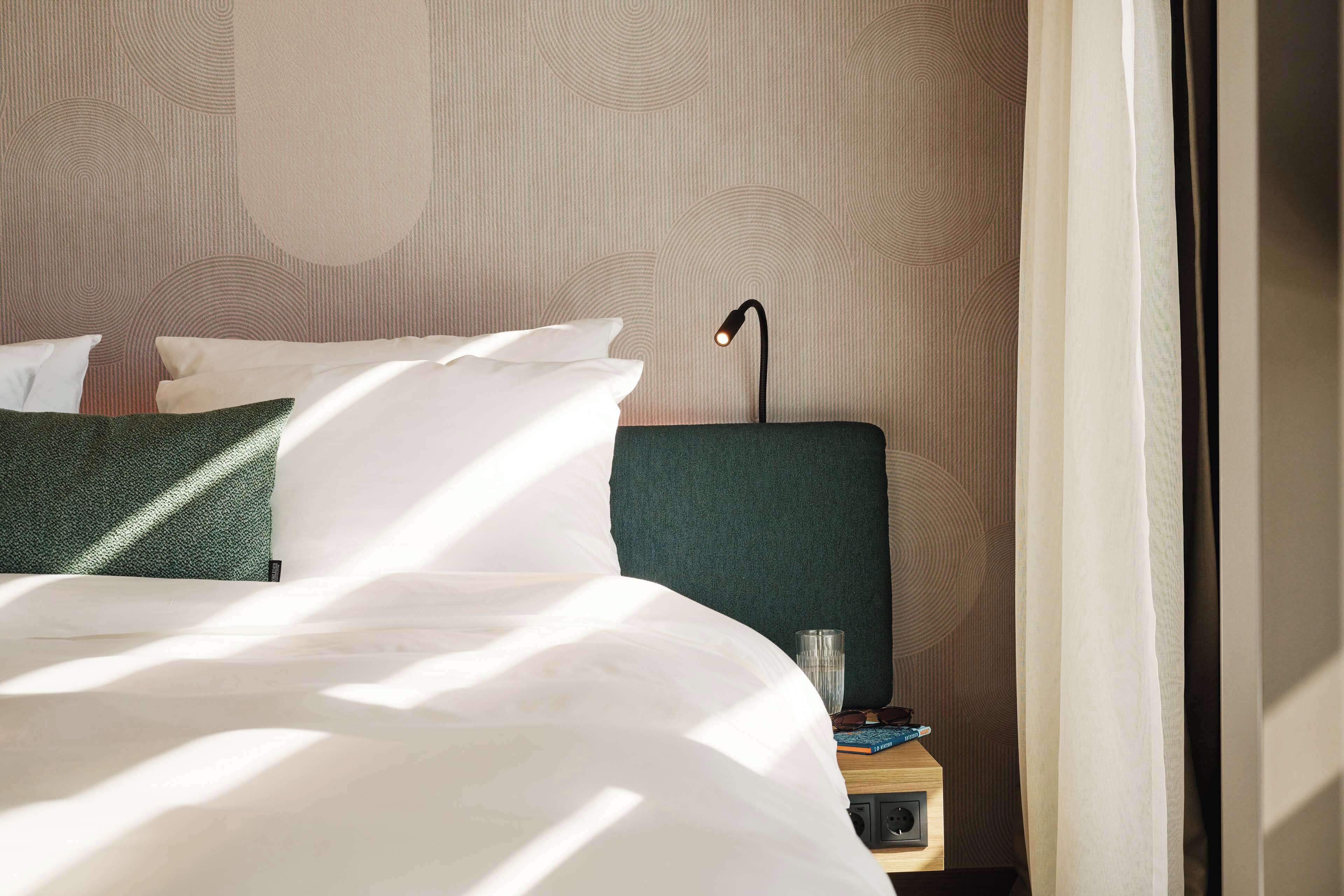 Un letto rifatto con cuscini bianchi e verdi, una testiera verde e un comodino con una lampada e un libro.