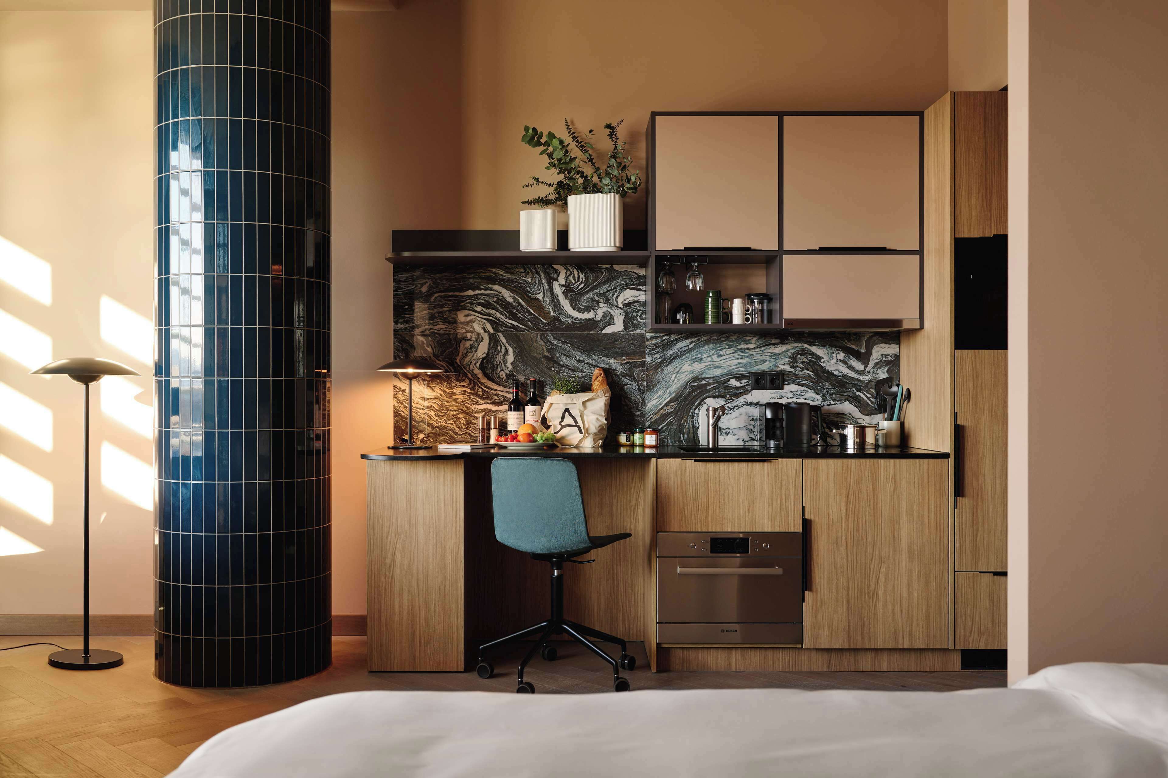 Une chambre moderne comprenant un lit, un bureau avec une chaise, une grande colonne cylindrique et un coin cuisine avec des placards et un pare-feu en marbre.