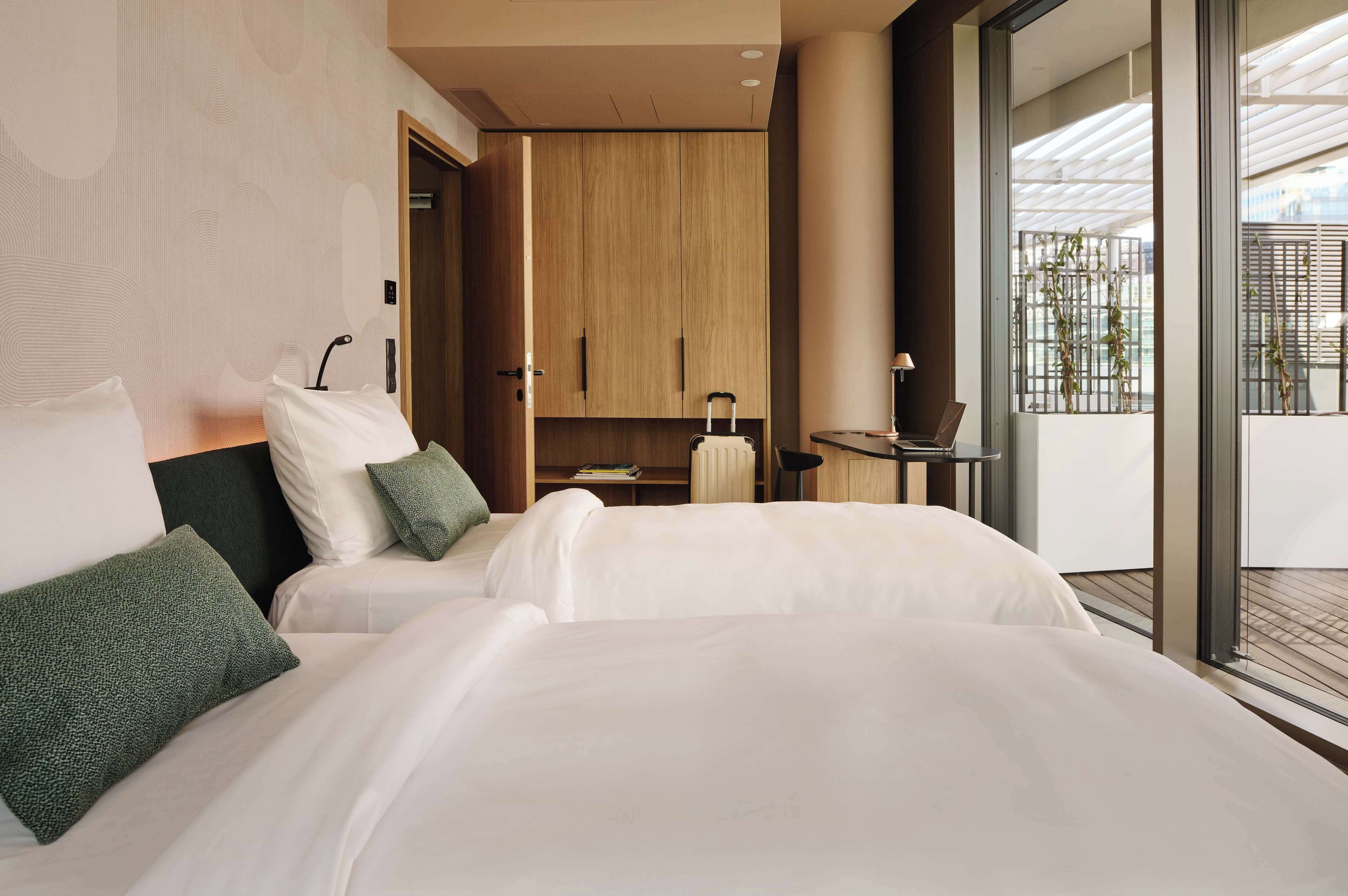 Habitación de hotel moderna con una cama grande, un armario de madera, una mesita con una lámpara y vistas al balcón exterior.