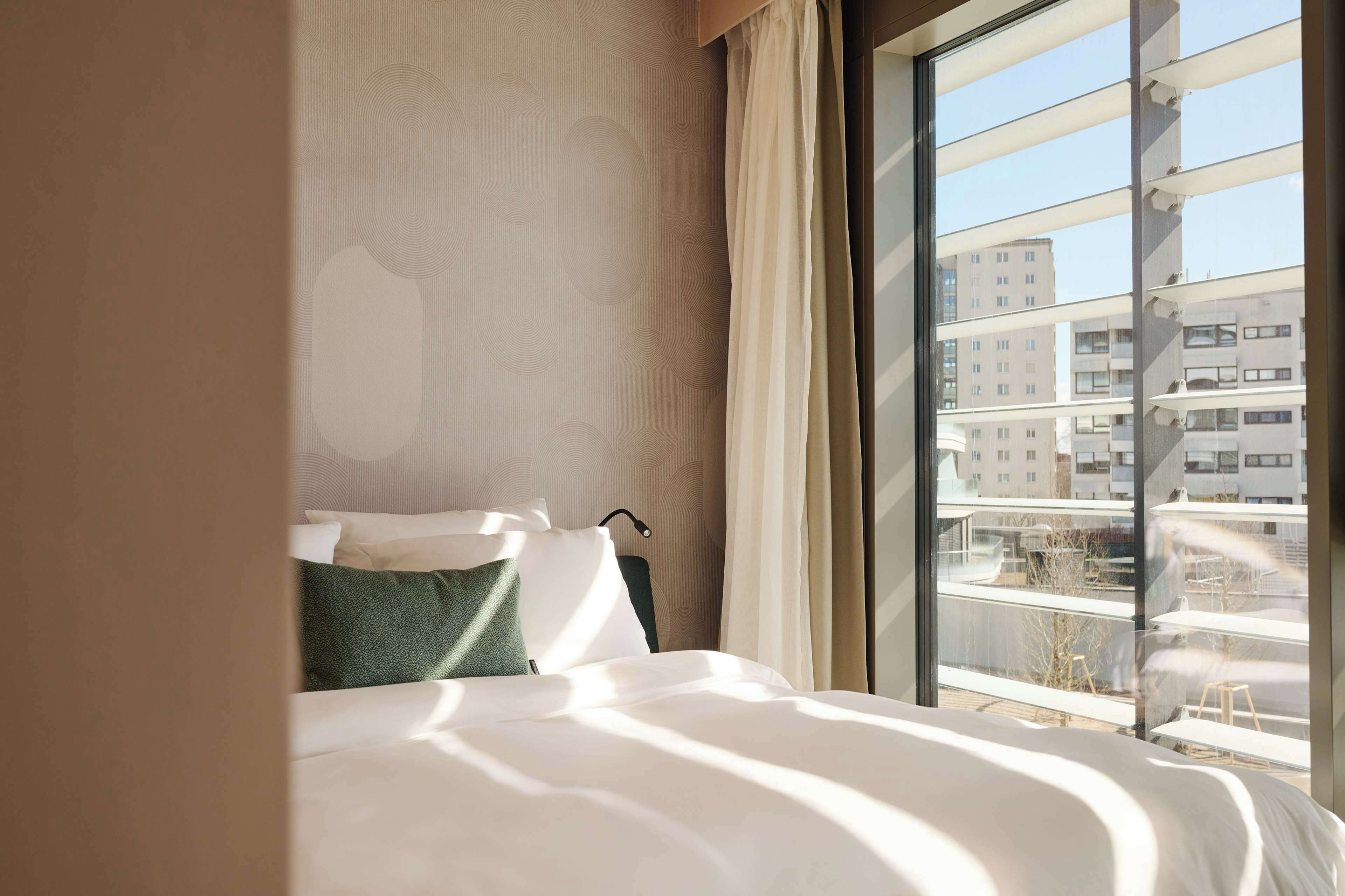 A hotel room with a large window, a bed, and a view of the cityscape.