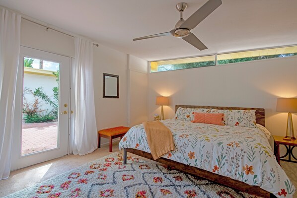 Un dormitorio con cama, ventilador de techo, mesita de noche, lámpara, ventana con cortinas y vistas al exterior.