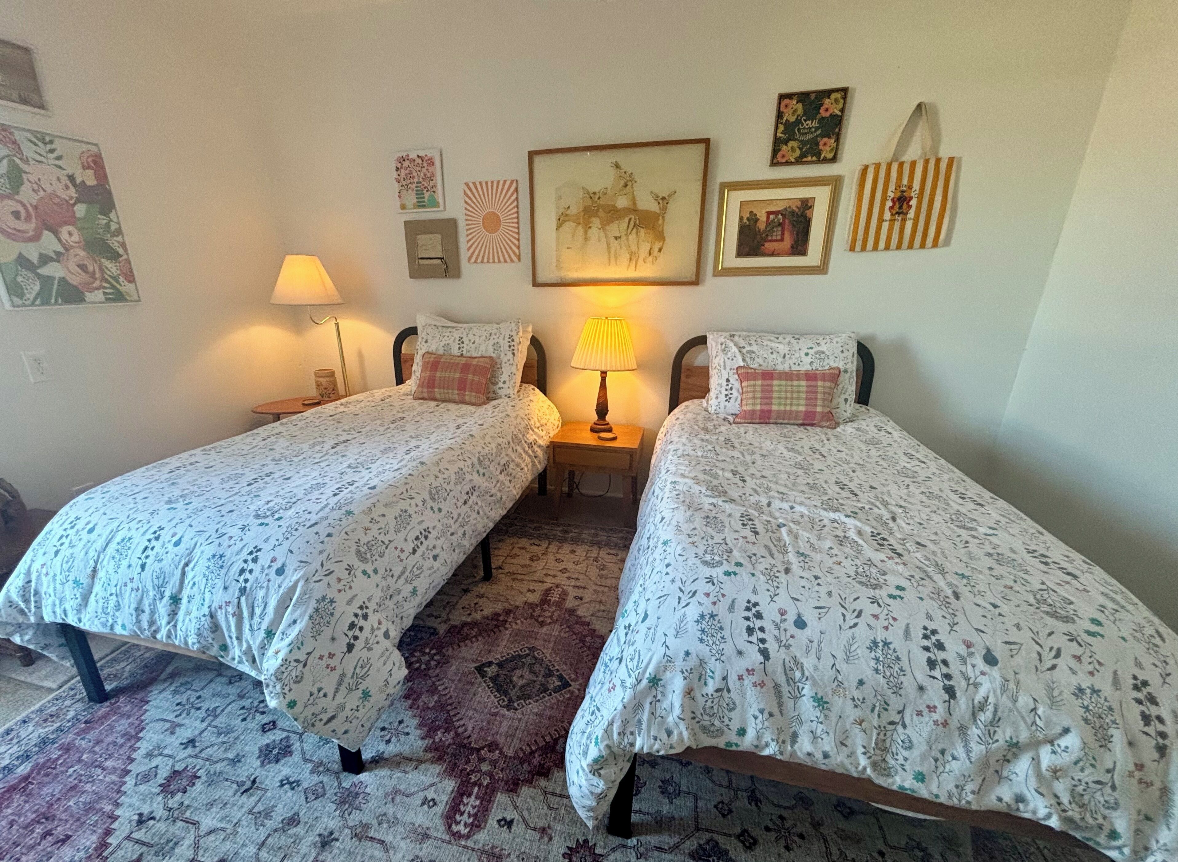 Ein Zimmer mit zwei Betten, jeweils mit blumigem Bettbezug, einem Teppich und Wanddekorationen, darunter gerahmte Bilder und ein Schild.