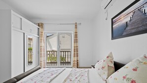 A bedroom with a bed, a window with curtains, a balcony, and a wall-mounted air conditioner.