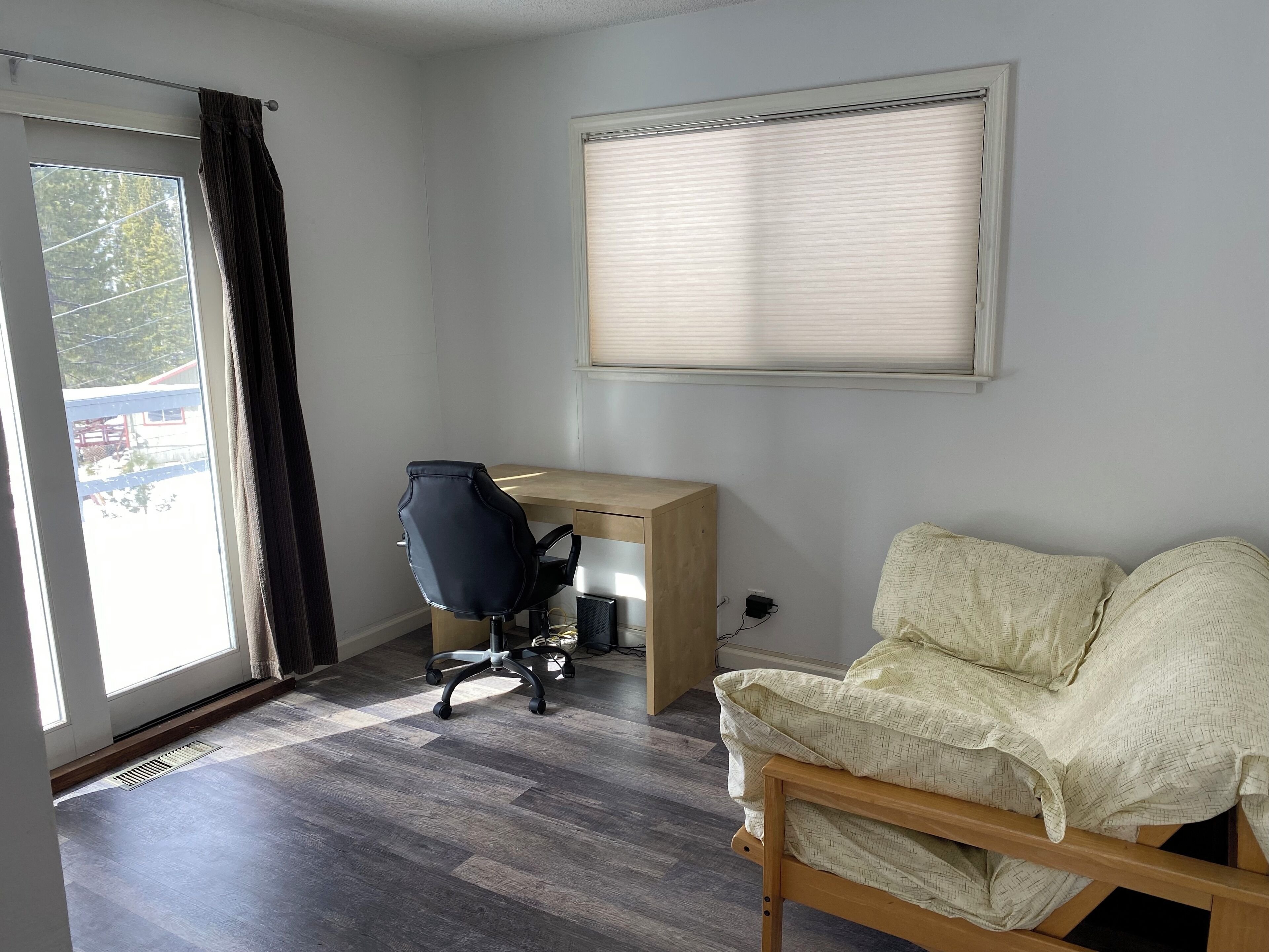 A room with a wooden desk, a black office chair, a beige sofa, and a window with blinds.