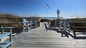 Une promenade en bois mène à des chaises et des tables installées sur la plage, avec un drapeau américain en arrière-plan.