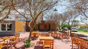 An outdoor seating area with wooden tables and benches, surrounded by trees and a building with a wooden facade.
