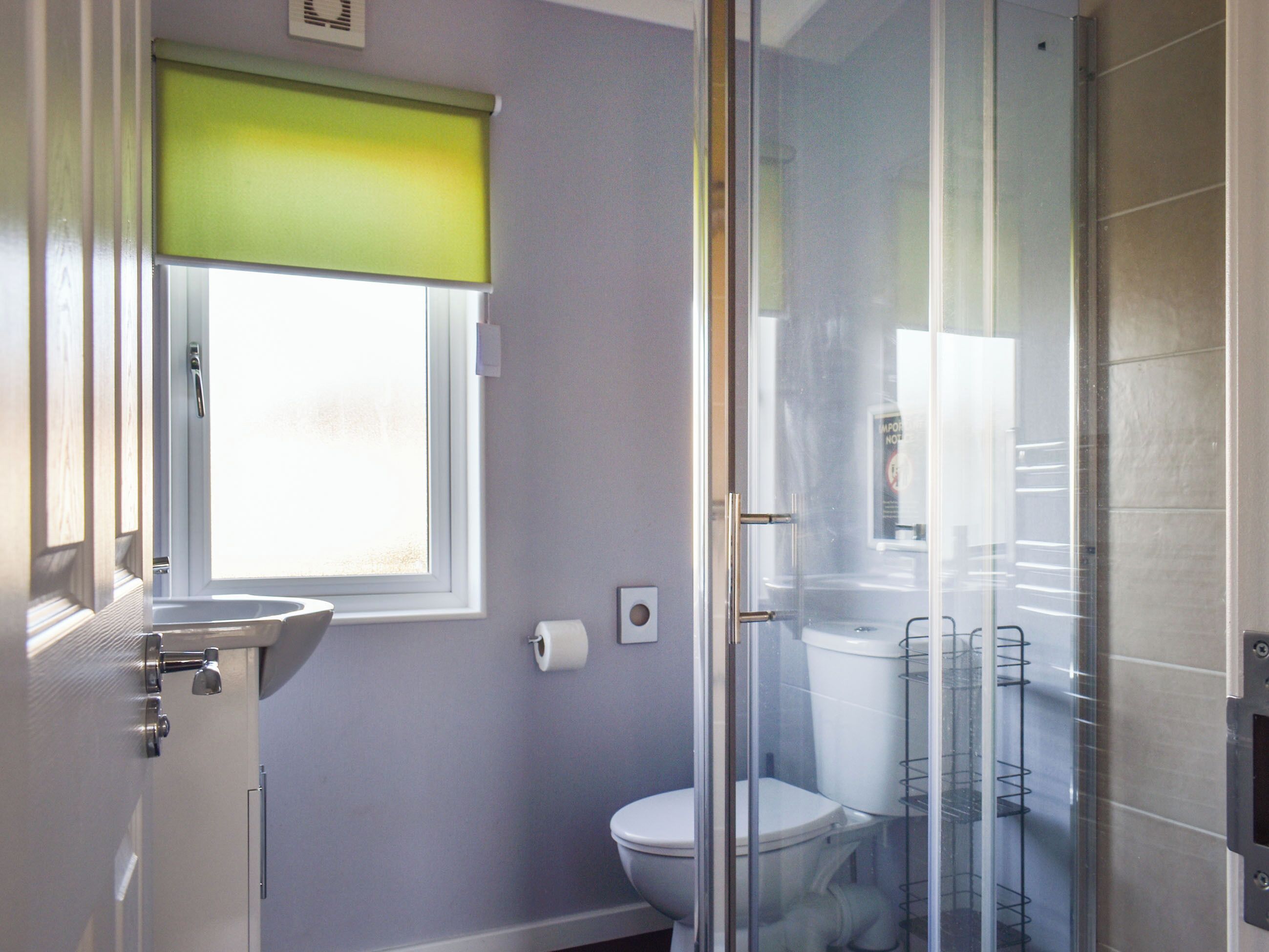 A bathroom with a toilet, a glass shower enclosure, and a window with a green blind.