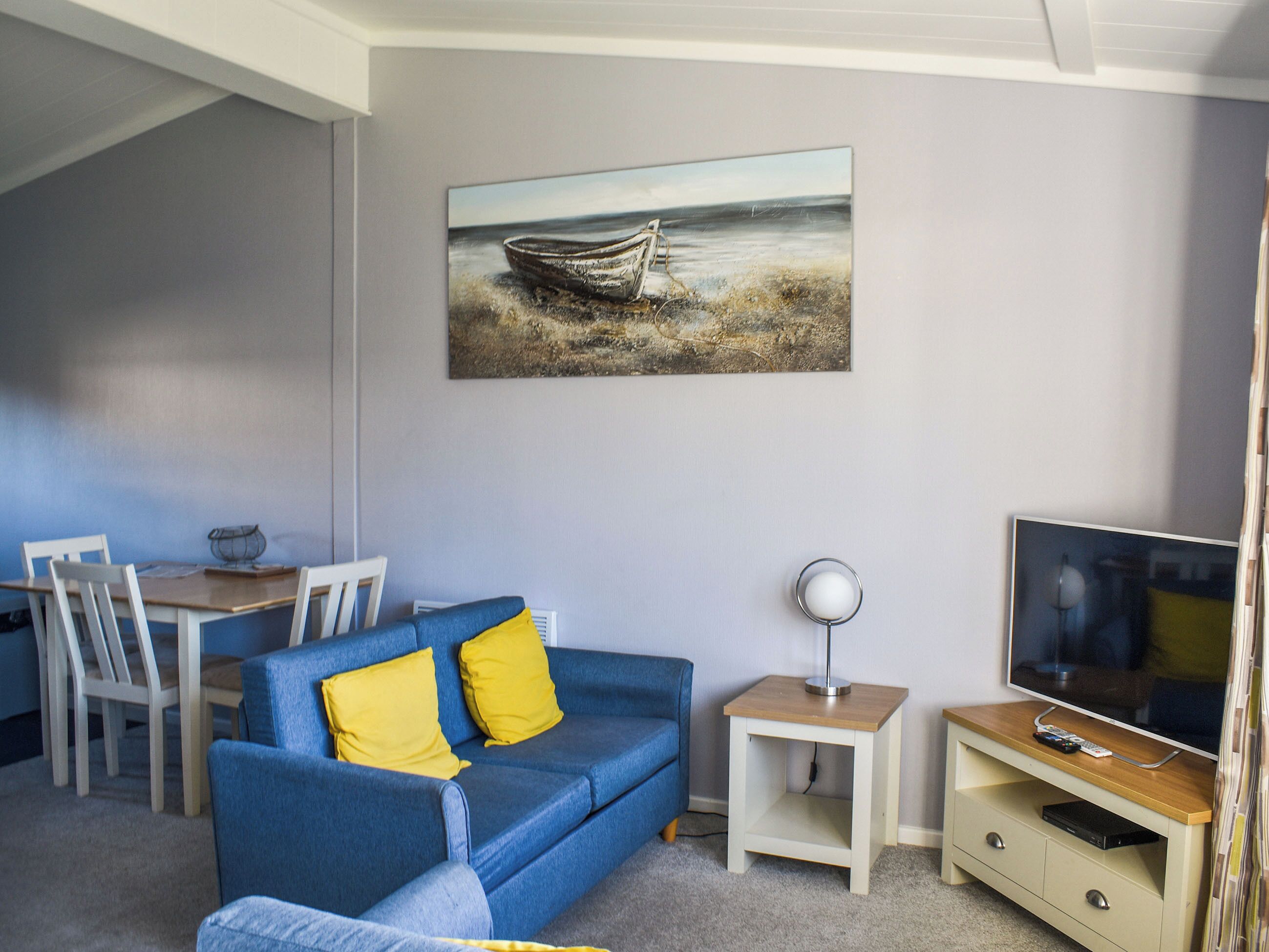A living room with a blue sofa, a painting of a boat on the wall, a TV, and a small table with a lamp.