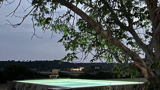 A rectangular pool with a wooden deck, surrounded by trees and a distant view of a hill with buildings. - Unity House Sleeps 4 with terrace - Masseria 1 by Rentbeat (Ceglie Messapica)