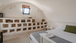 Un dormitori amb sostre inclinat, un llit amb edredó gris, una tauleta de nit amb una làmpada blanca i una petita finestra amb persianes de fusta.