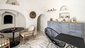 Una habitación con una mesa redonda, dos sillas de mimbre, una silla de metal negro, un armario azul con estantes y un suelo de piedra.